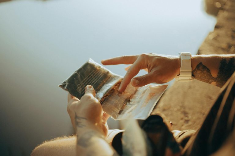 A close-up of tattooed hands holding a plastic bag near a calm body of water in sunlight.