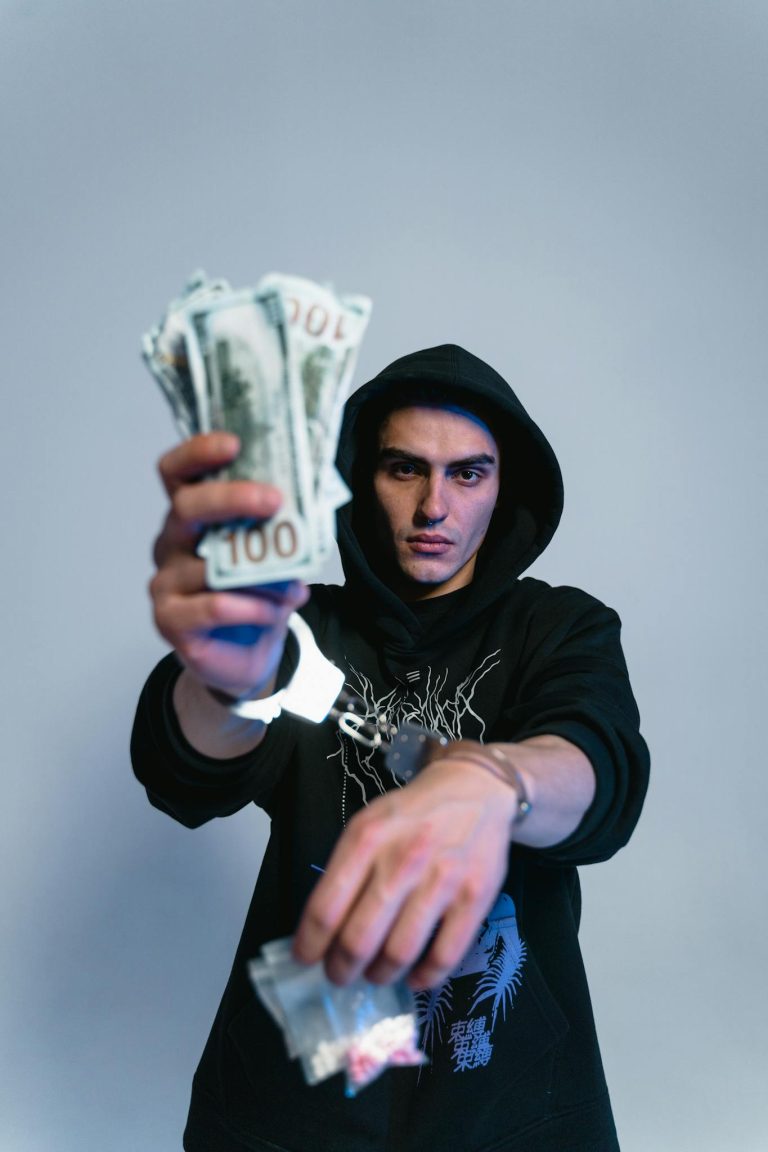 A young man in a dark hoodie holds cash and small bags while handcuffed, conveying a criminal theme.
