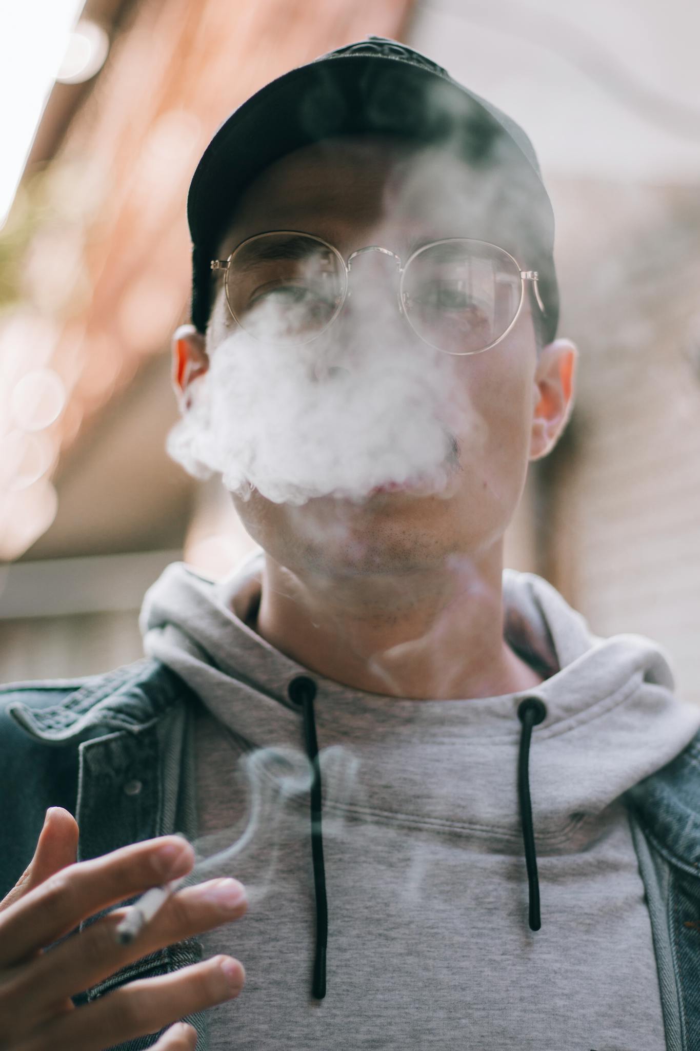 A young man in a hoodie and cap smokes outdoors, exhaling vapor.