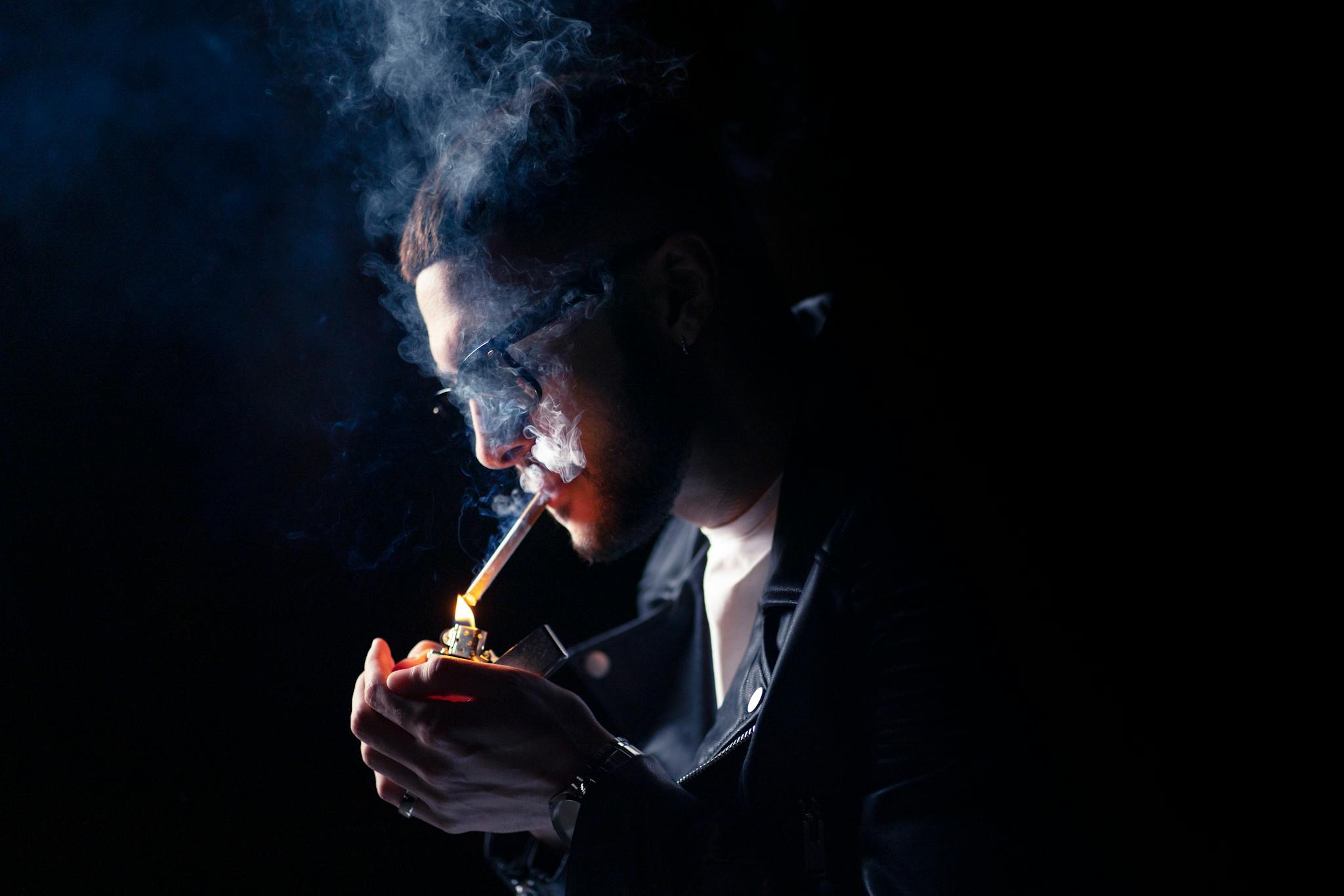 A young man in a leather jacket lighting a cigarette, surrounded by smoke.